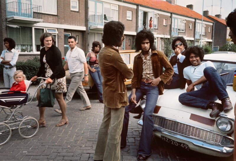 A group of young men with 1970s hairstyles and clothing gather around and sit on a white car parked on a residential street, while people, including a woman with a pram, pass by in the background.
