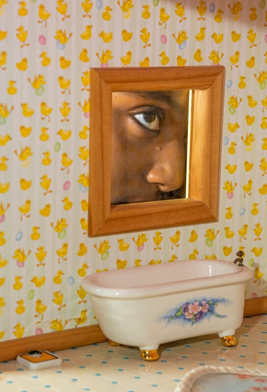 A close-up of a person's face reflected in a small mirror above a mini bathtub with floral decoration, set against wallpaper with yellow duck illustrations.