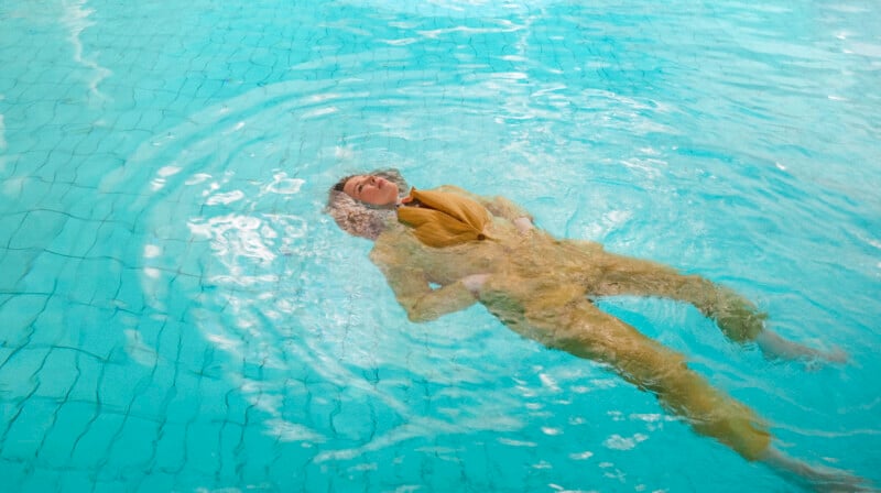 A person in a yellow outfit floats on their back in a swimming pool, with water rippling around them and their face partially above the water’s surface.