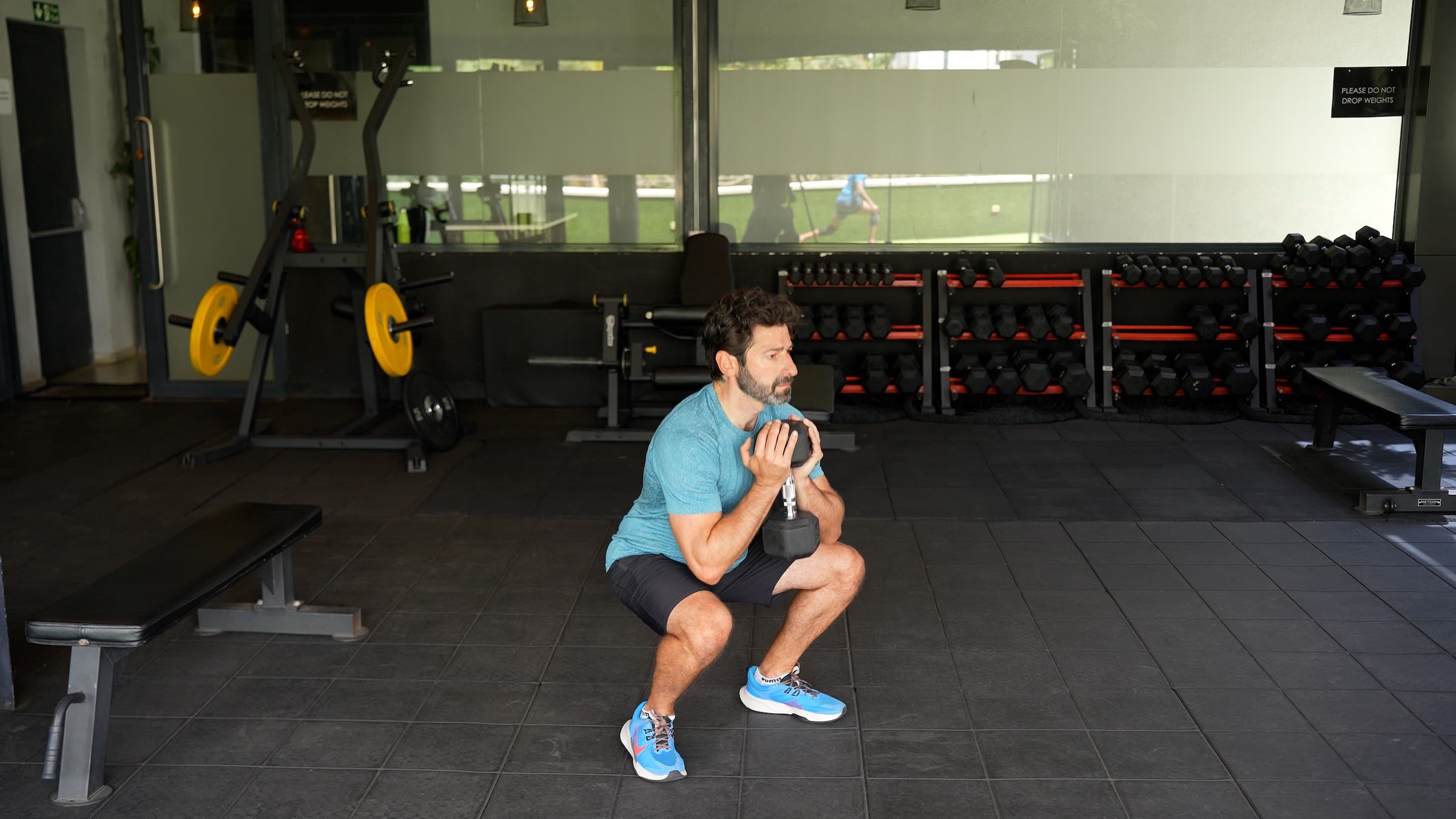 a man with dark hair wearing a blue shirt and black shorts does a goblet squat in a gym holding a weight at his chest