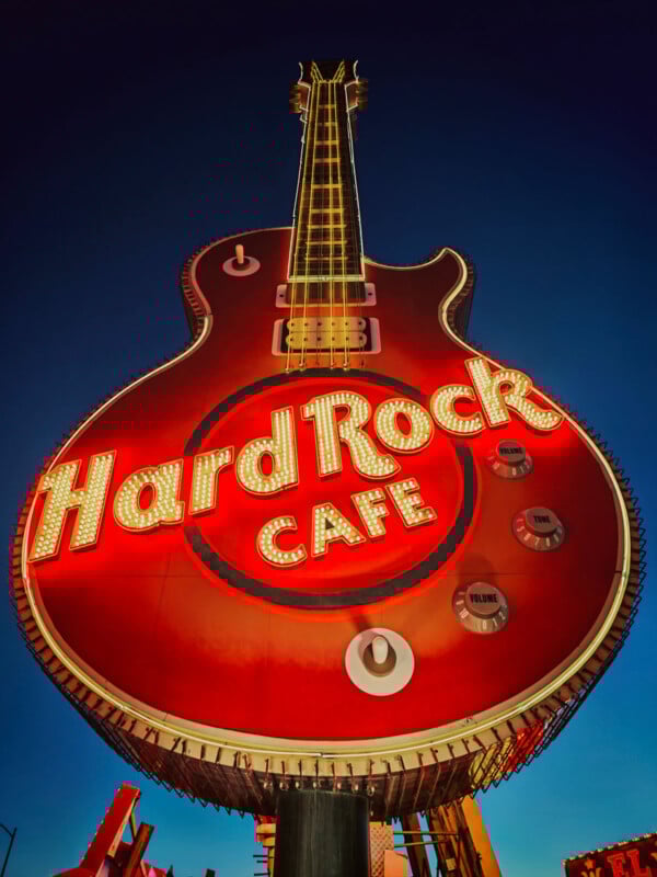 Large neon Hard Rock Cafe sign shaped like a red electric guitar, glowing brightly against a deep blue evening sky. The sign features illuminated letters and guitar details like strings and control knobs.