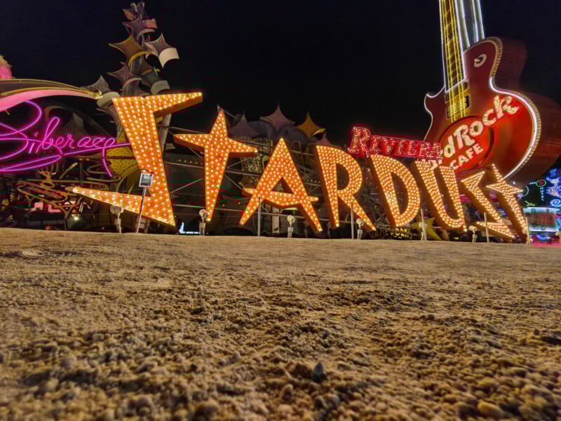 Illuminated vintage "Stardust" sign with large marquee lights, set against a night sky. Other neon signs, including a guitar and "Liberace," are visible in the background. The ground is sandy and textured.
