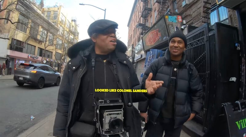 Two people stand on a city sidewalk, smiling and talking. One holds a large camera and wears a hat and coat. The other wears a beanie and puffer jacket. Text on screen reads, "LOOKED LIKE COLONEL SANDERS." Urban buildings line the street.
