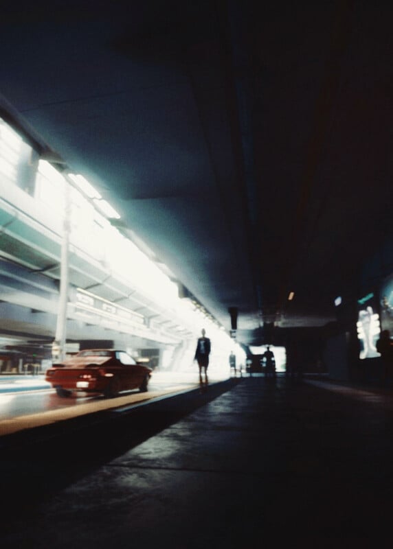 A blurry, dimly lit train station platform with a red car on the left and a silhouetted person walking toward a bright, modern structure in the background. The atmosphere is moody and futuristic.