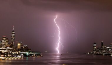 Lightning forms: A jagged, glowing, vertical white line striking the water between two areas of tall buildings.