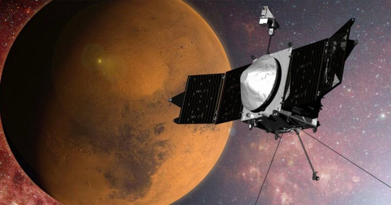 A spacecraft with solar panels orbits close to the reddish surface of Mars, set against a backdrop of space filled with stars and galaxies.