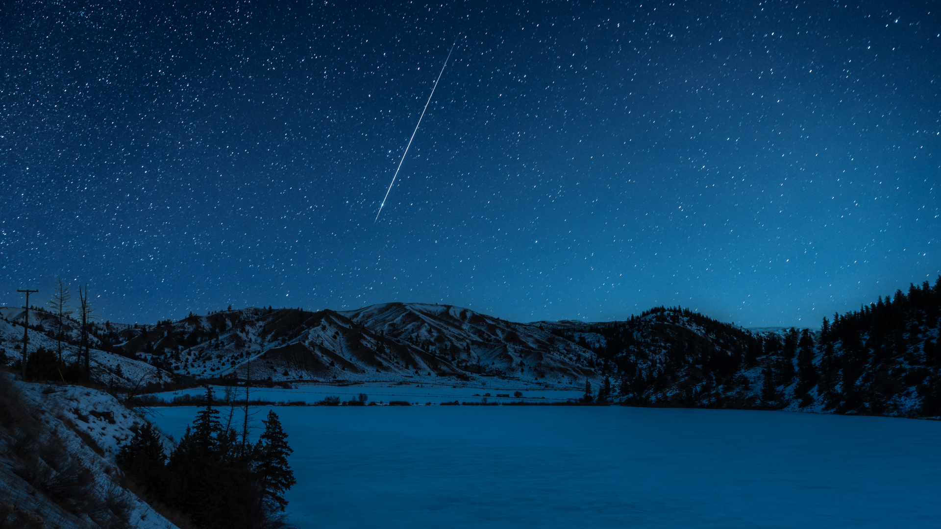 A meteor is pictured streaking earthwards through a starry night sky above a frozen lake bordered by snowy hills and evergreen trees.