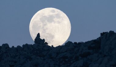 A full moon is photographed rising above a rocky hillside in the dark blue evening sky.