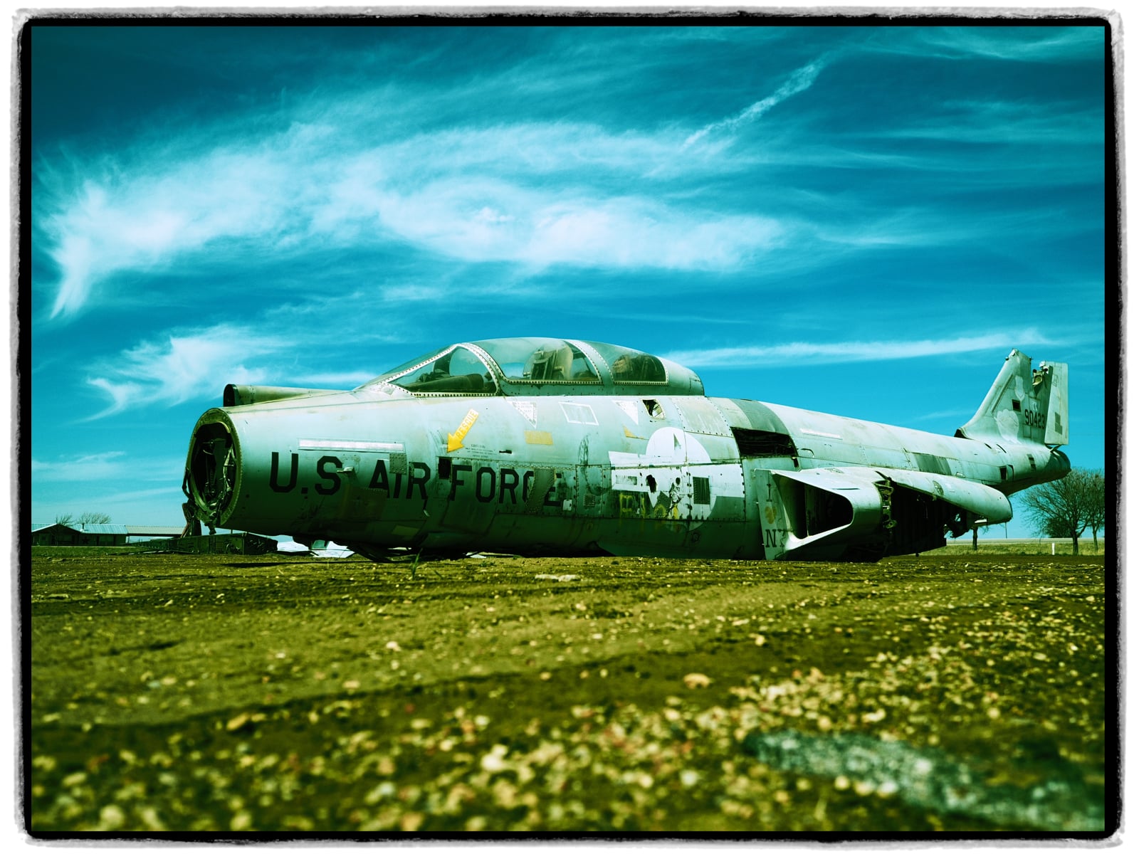An old, weathered U.S. Air Force jet sits abandoned on a dirt field under a bright blue sky with wispy clouds. The aircraft shows signs of rust and faded paint, surrounded by sparse grass and distant trees.