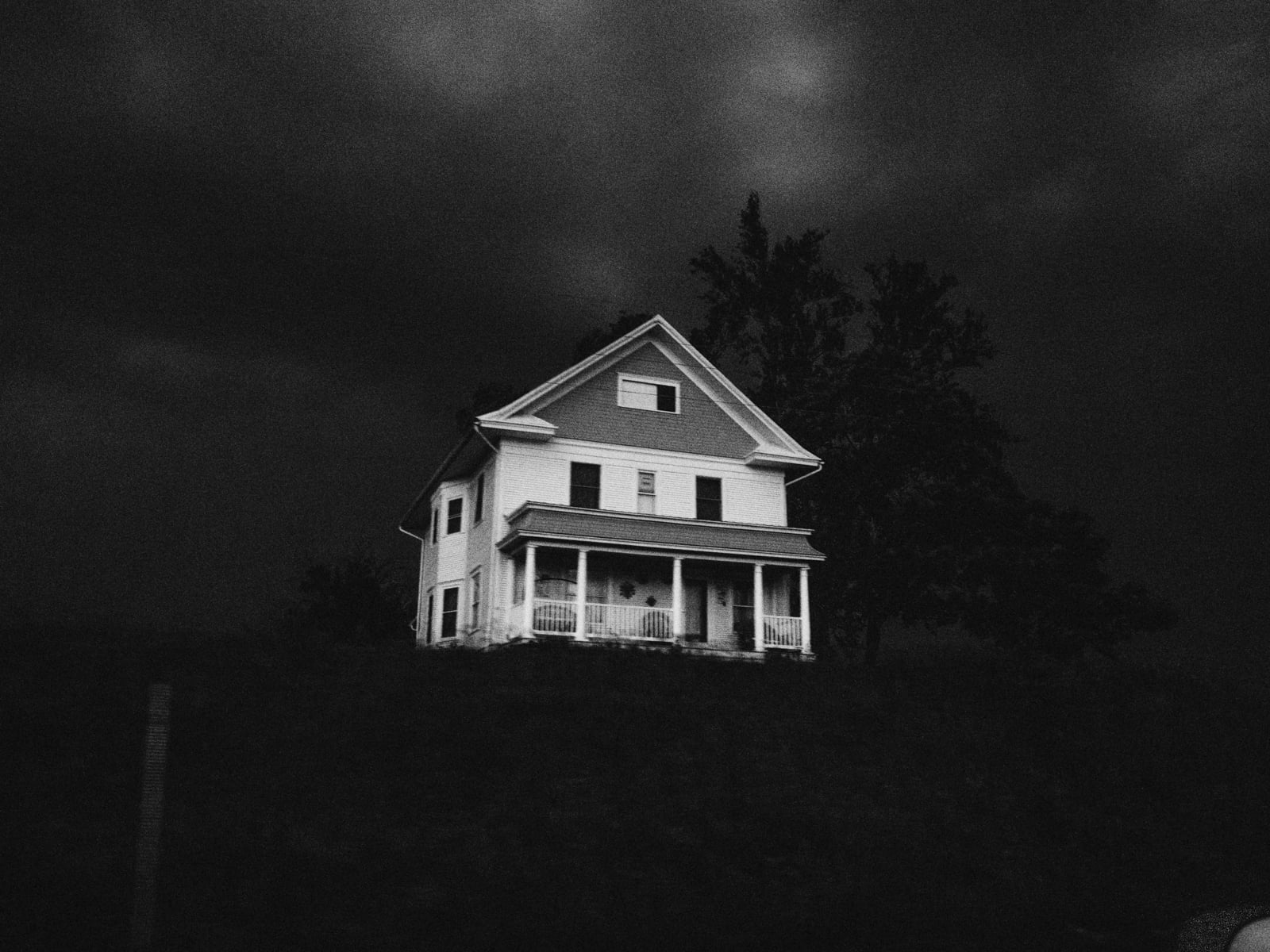 A white, two-story house with a porch stands alone on a hill under a dark, cloudy sky, creating a moody and eerie atmosphere.