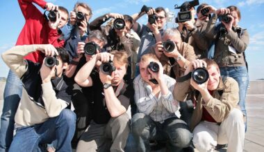 A group of photographers and videographers crouch together outdoors, all aiming their cameras and video recorders directly at the viewer under a bright, clear sky.
