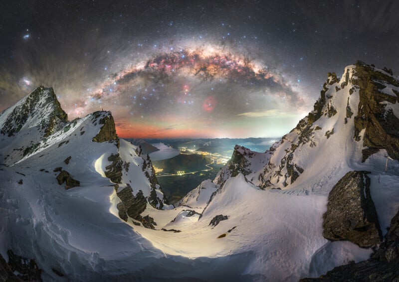 A starry night sky with the Milky Way arched above snowy mountain peaks, glowing lights from a valley town below, and rugged rocks framing the scene.