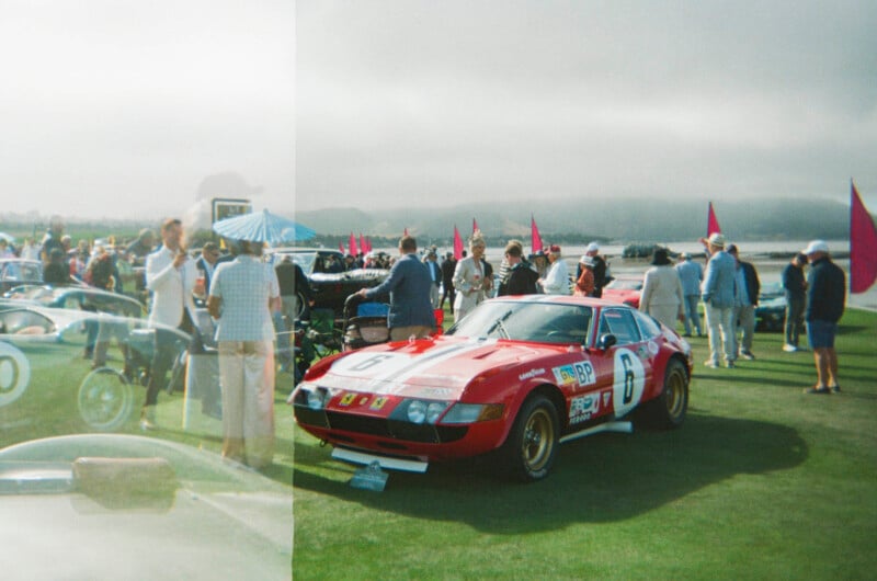 A red vintage race car with the number 6 is parked on a grassy field surrounded by people and other classic cars at an outdoor car show under a cloudy sky.