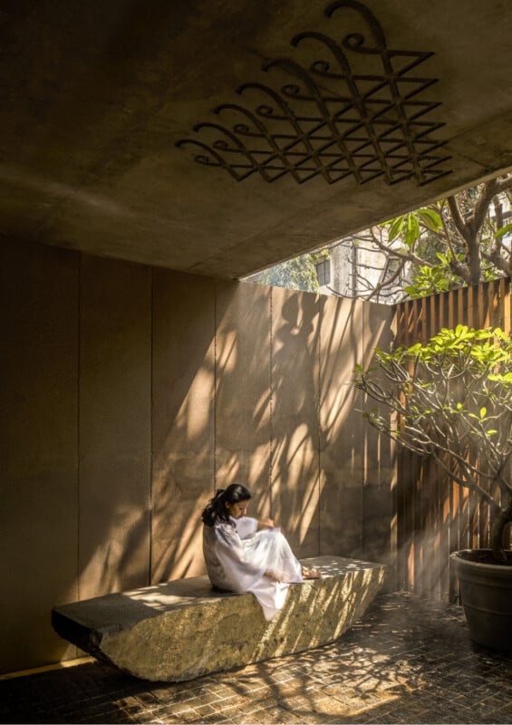 A woman in a white dress sits on a stone bench in a sunlit, modern outdoor space with textured walls, a geometric ceiling design, and a tree casting patterned shadows.
