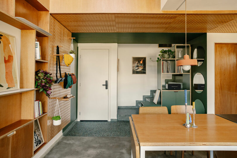 Modern interior with wooden shelves, colorful wall hooks, a green accent wall, vinyl records, a dining table, and minimalist decor. A white door and small staircase are visible in the background.