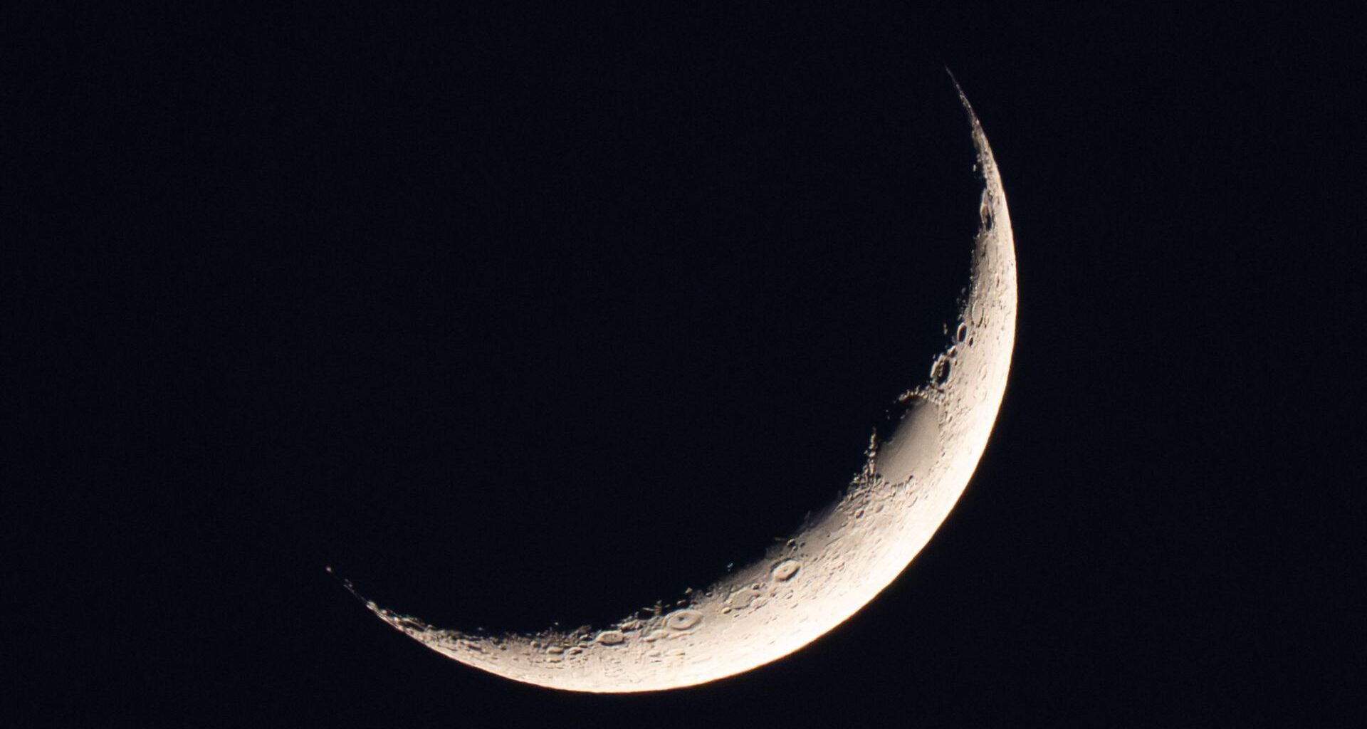 A photo of a thin waxing crescent moon shining in a black night sky. A dark oval is visible on the upper section of its sickle-like profile. Elsewhere, large craters can be seen pockmarking the illuminated wedge of the lunar disk.