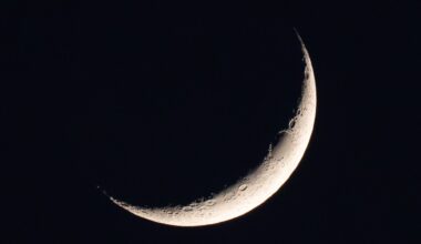 A photo of a thin waxing crescent moon shining in a black night sky. A dark oval is visible on the upper section of its sickle-like profile. Elsewhere, large craters can be seen pockmarking the illuminated wedge of the lunar disk.