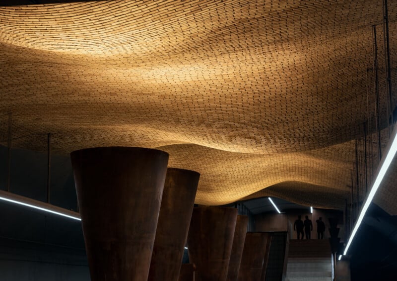 Golden, wavy brick ceiling with large, rust-colored cylindrical columns below; four silhouetted people stand on stairs in the distance under modern, angular lighting.