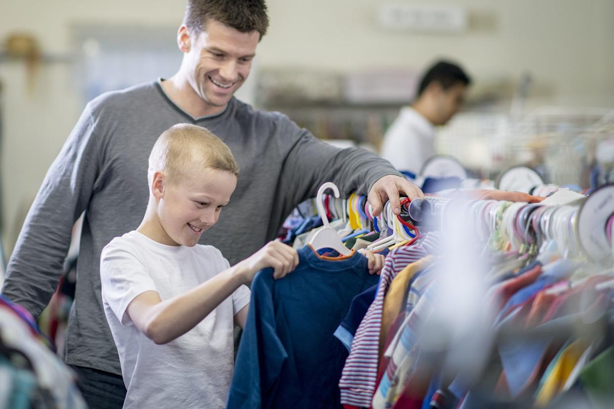 A little boy and his father are looking through a rack of clothes while shopping together. The boy has pulled out a long sleeve shirt to buy.
