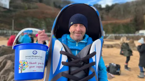 BBC Man wearing shoe costume on beach holding bucket collecting money for Jersey Hospice Care