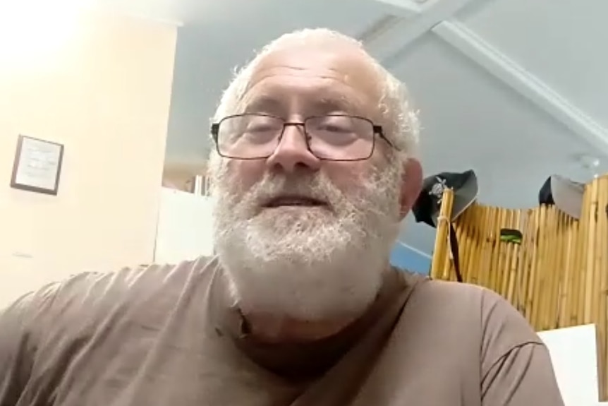 A man with a white beard wears a brown shirt and glasses inside a small room.