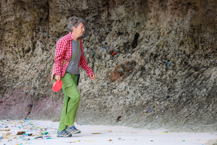 A woman wearing green pants and a pink shirt walks on the beach next to a rock with coloured pieces of plastic dotted around it.
