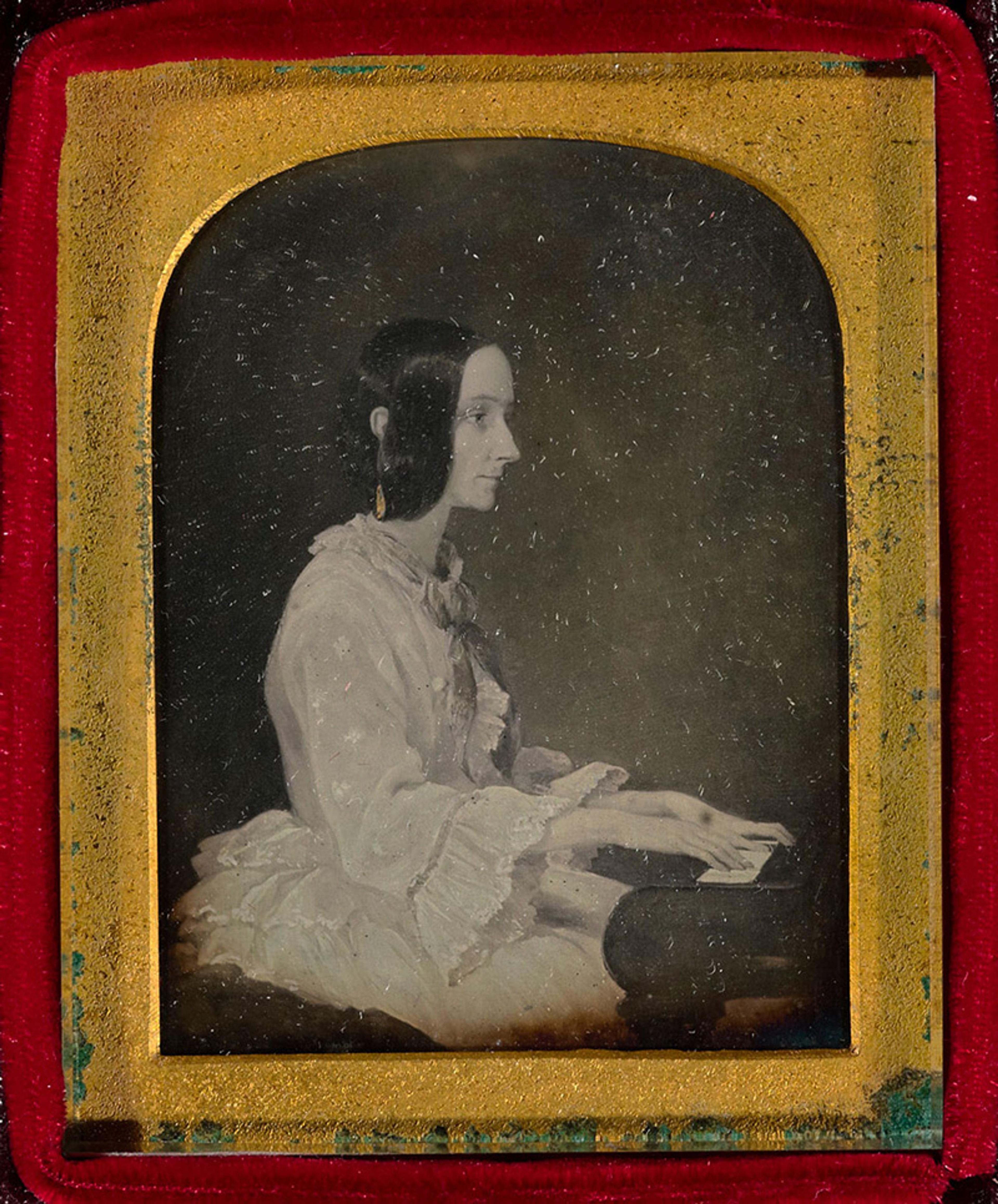 Daguerreotype of a woman in profile playing the piano, wearing a ruffled dress, set in a red and gold frame.