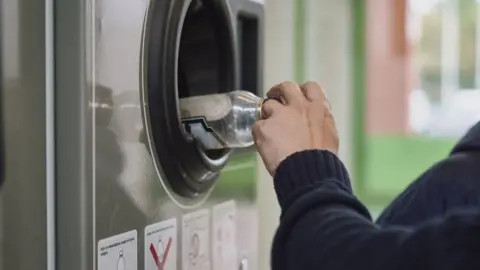 Getty Images A glass bottle is being placed into bottle return machine. A person is holding the top to the bottle with their left hand, with their arm, covered by a dark jumper, visible. The machine is grey, with the bottle entering a round black hole. 