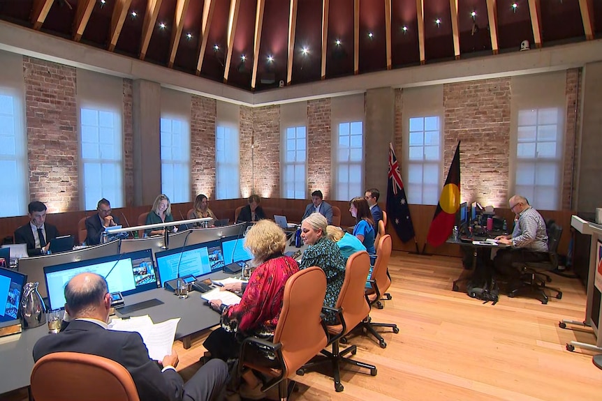 A large long meeting table with councillors sitting at it inside a high-ceiling building.