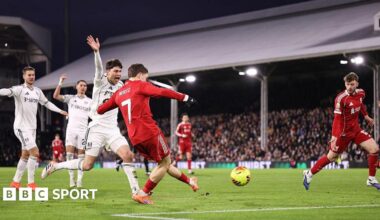 Liverpool's Florian Wirtz scores against Fulham. He is in the penalty area and has struck a yellow ball with his left foot. Liverpool's kit is red and he is wearing orange boots. A Fulham player, wearing all white, is next to him and is raising his right arm to call for offside. Several other players can be seen in the background along with thousands of fans in one of the stands.