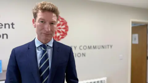 A man with short ginger hair looking at the camera. He is wearing a blue suit blazer, a blue tie with green stripes and a blue shirt. You can see part of the Guernsey Community Foundation logo behind him