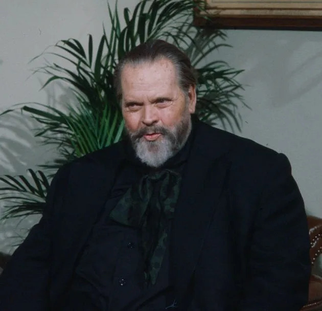 A distinguished older man with a beard and mustache sits in a room with framed art and a plant, wearing a dark suit