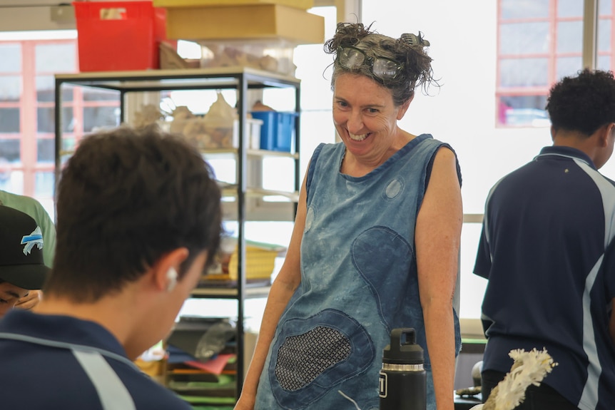A woman wearing a blue singlet and glasses on her head smiles and leans on a table inside a room with students.