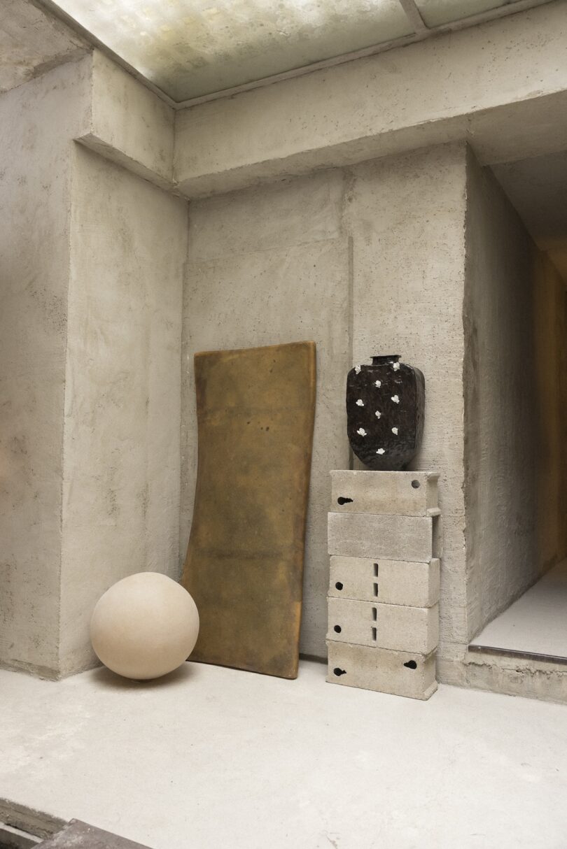 A beige sphere, a rectangular slab, and a dark vase with white spots rest on stacked concrete blocks against a rough, light-colored wall in a minimalist interior space.
