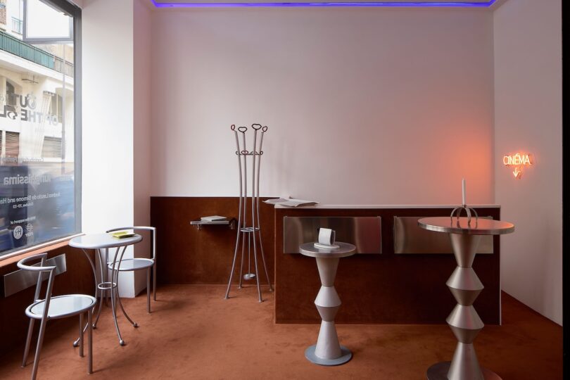 Modern room with geometric metal furniture, a round table with two chairs, a counter, abstract sculptures, coat rack, and a neon "CINEMA" sign on a brown carpeted floor.
