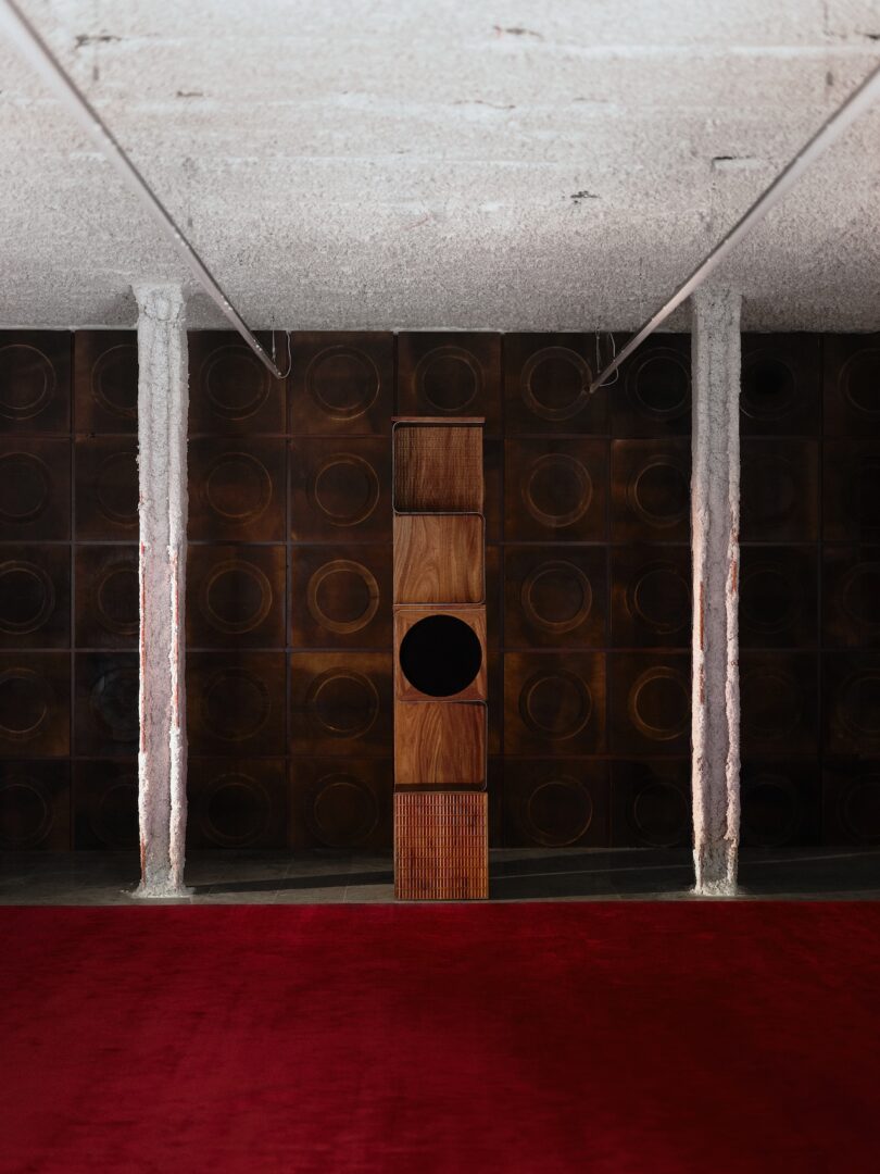 A tall wooden speaker stands centered against a dark, textured wall with circular patterns; white pillars and a red carpet are in the foreground.