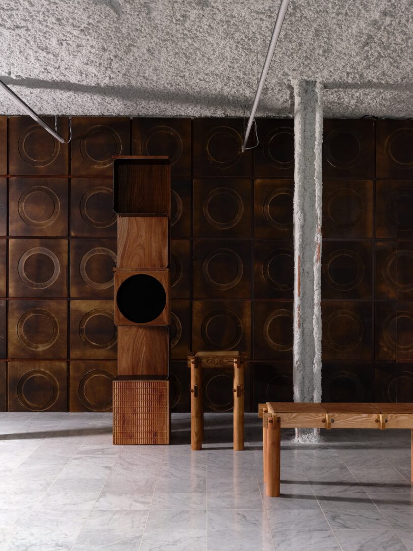 A minimalist interior features textured brown wall panels with circular patterns, wooden geometric furniture, a marble floor, and an exposed concrete column.