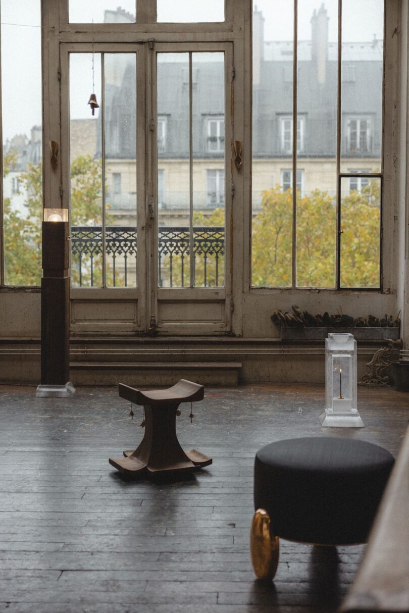 A wooden stool, black ottoman, and two pillar candle holders on a dark wooden floor near tall windows with a view of buildings and trees outside.