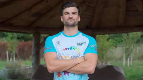 Joe Dilling Joe Dilling poses for the camera with his arms crossed in a outdoor area and trees in the background. He is wearing a charity running top.