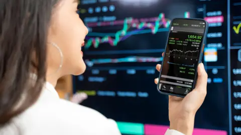 Getty Images A woman in a white shirt looks at a line graph on her phone, with a big black stock market trading screen in the background.