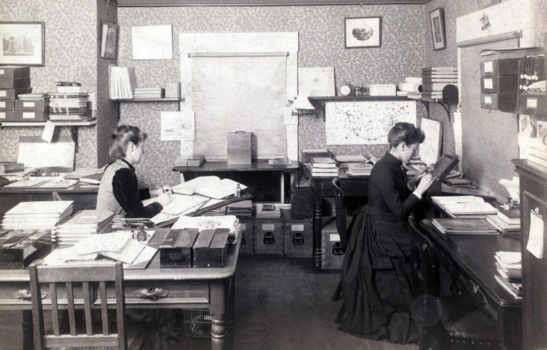 Black and white photo of two women in late-nineteenth-century clothing in an office, processing astronomical data on paper.