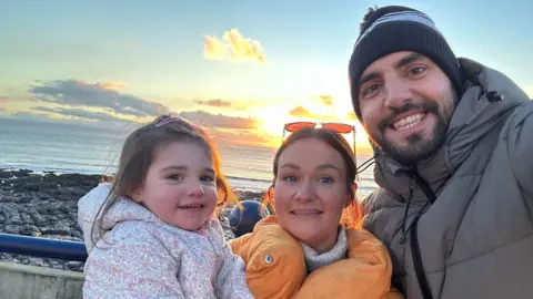 Ellie James A woman and her husband pose with their young daughter, all wearing coats, at the seaside.