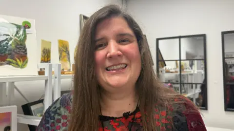 A woman with long brown hair is smiling and wearing a red and blue dress as she stands in an art gallery with various artworks on white shelves in front of white walls and mirrors with black frames.