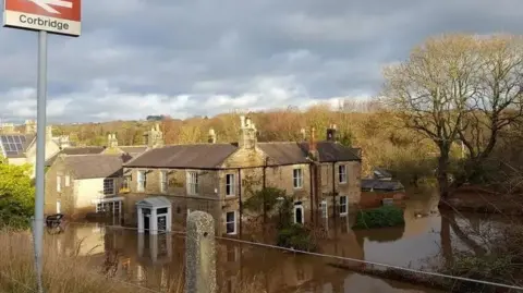 Flood water comes up to the bottom third of the door of a stone pub. A sign for Corbridge Station is in the foreground.