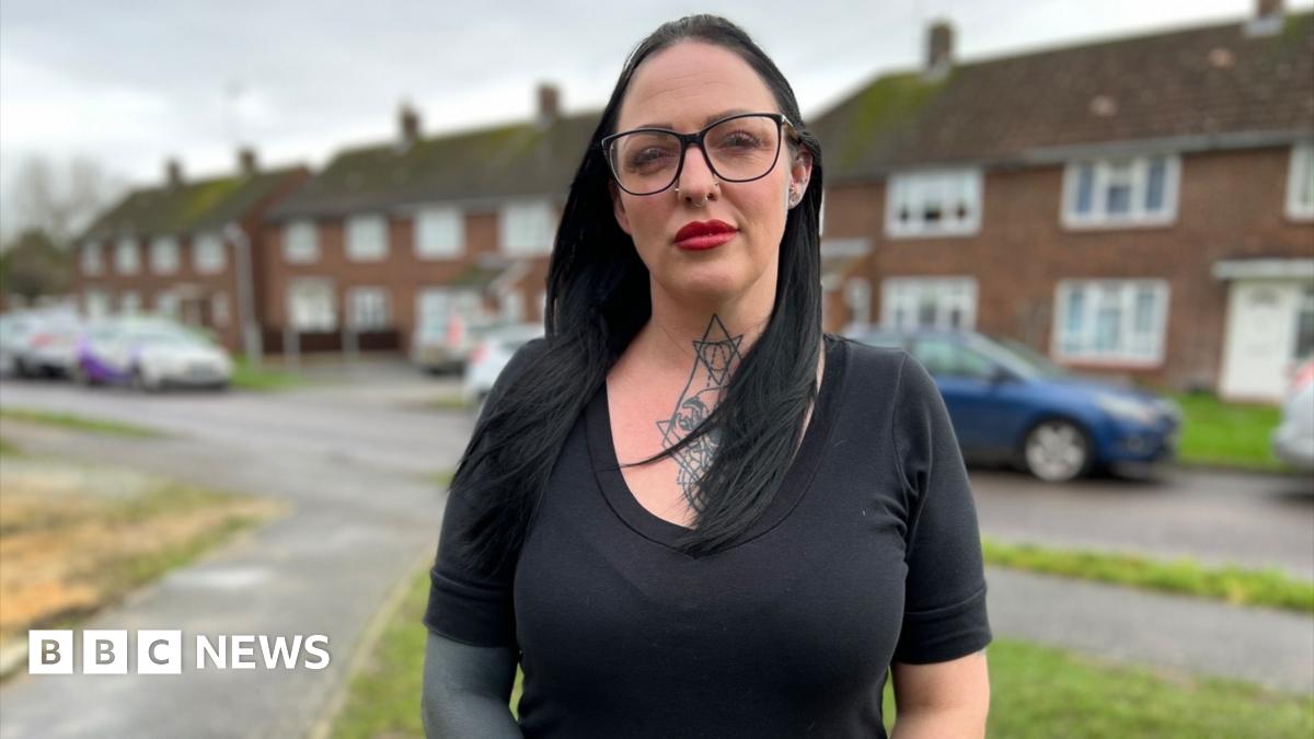 Sam Cook stands in front of a row of houses with a road, parked cars and grass verge in front. Sam has chest-length black hair. She wears a black T-shirt and has black-rimed glasses, as well as red lipstick. Sam has a large tattoo on her neck and a sleeve tattoo on her right arm.