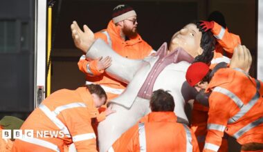 Five men in high viz orange jackets, carrying one of Beryl Cooks sculptures out of a van. The sculpture is of 'Dancing Tom'.