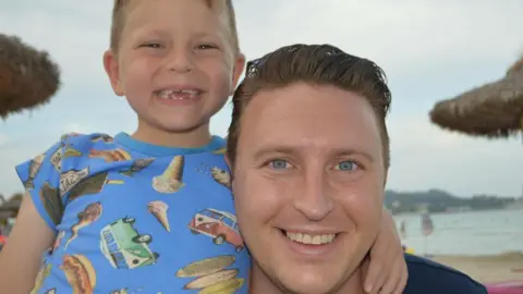 Tim Sadler Image of a young boy with a blue t-shirt with images of camper vans and ice creams with his arm around a man with blue eyes and short brown hair.  The boy has gaps in his teeth but he and the man are smiling at the camera. In the background can be seen sand, sea and beach umbrellas.