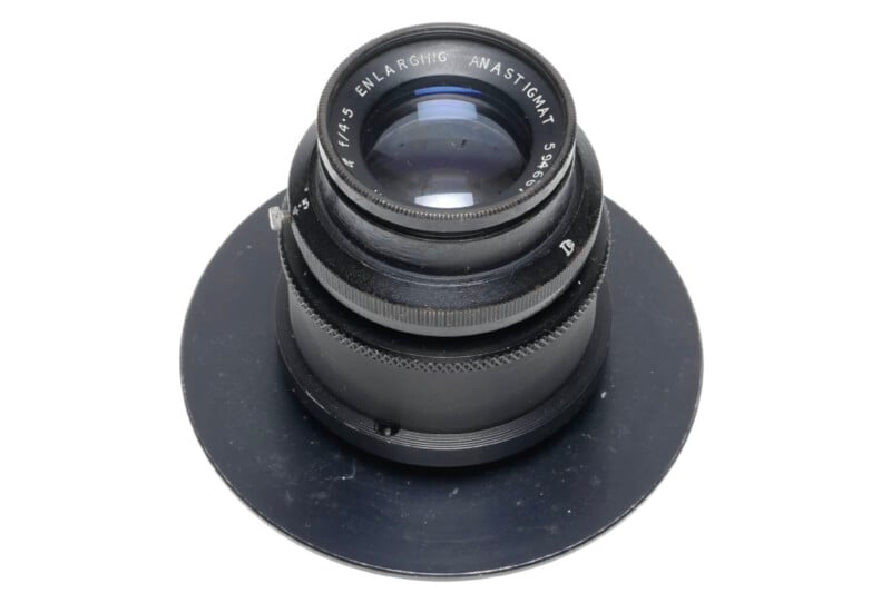 A black camera lens labeled “ENLARGING ANASTIGMAT f:4.5” is mounted on a round metal plate, viewed from above on a white background.