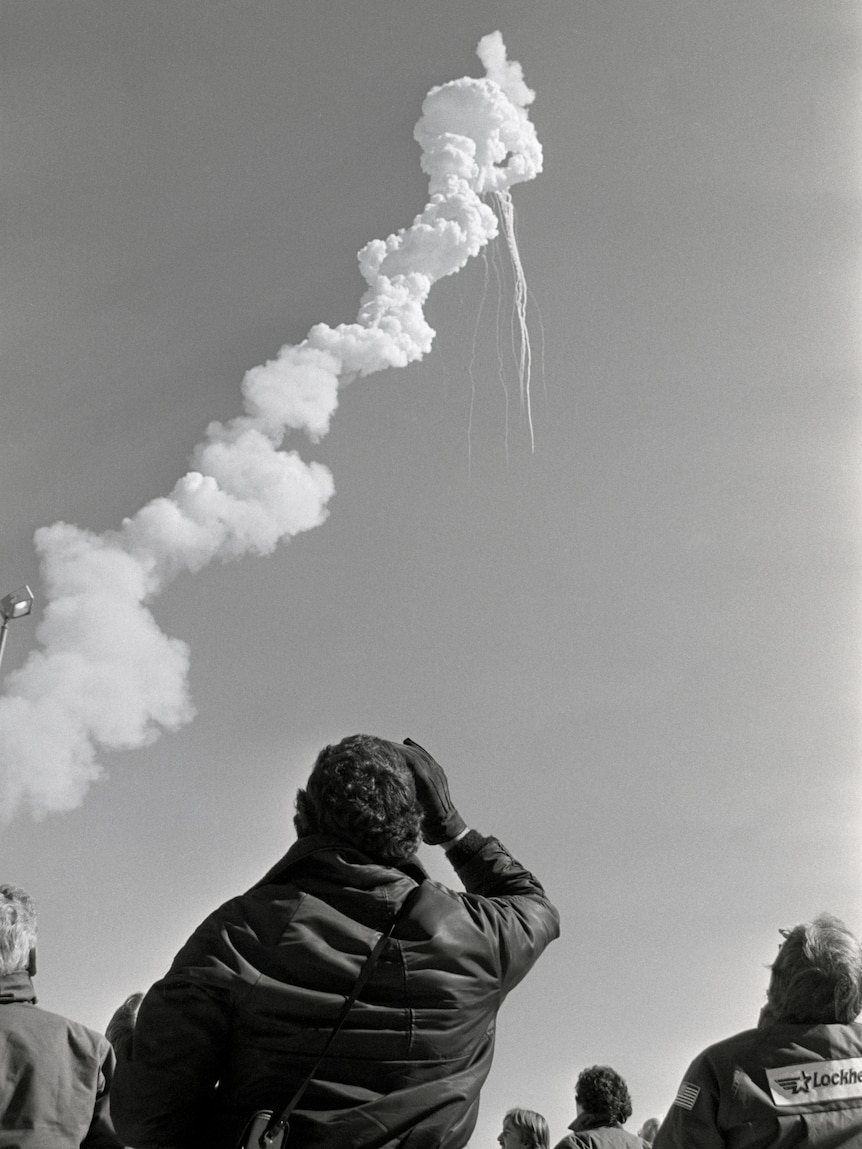 Black-and-white photo of the backs of several rugged-up onlookers peering up at a twisted cloud with downward streaks.