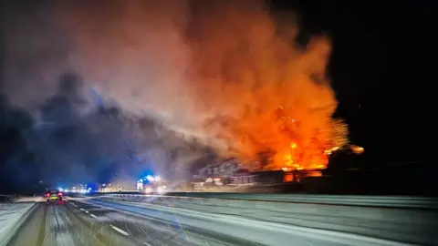 Hazel Robbie A snowy road with a large fire engulfing houses close to the carriageway with orange flames and smoke